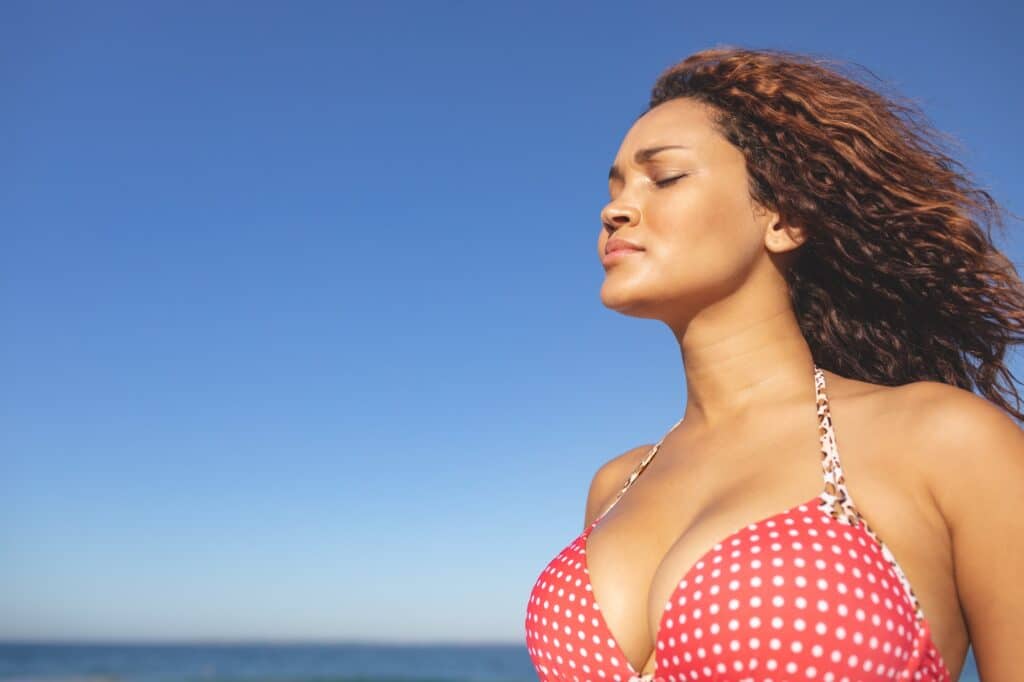 Side view of beautiful mixed race woman in bikini standing with eyes closed on the beach